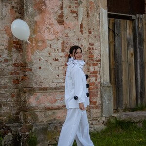 May include: A person in a white clown costume with a ruffled collar and black pom-poms, holding a white balloon. The costume includes a white jacket and pants. The background features a weathered brick wall and wooden door.