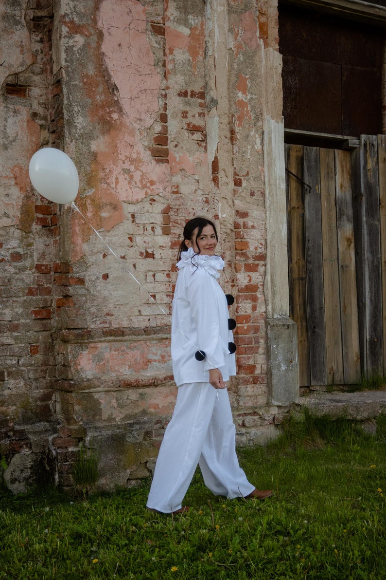 May include: A person in a white clown costume with a ruffled collar and black pom-poms, holding a white balloon. The costume includes a white jacket and pants. The background features a weathered brick wall and wooden door.