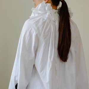 May include: A white, long-sleeved shirt with a ruffled collar. The shirt has a loose fit and is made of a lightweight fabric. The sleeves end at the elbow. The model has long, dark brown hair tied in a ponytail.