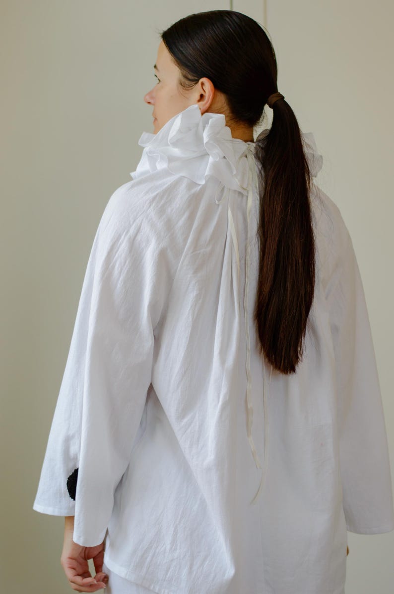 May include: A white, long-sleeved shirt with a ruffled collar. The shirt has a loose fit and is made of a lightweight fabric. The sleeves end at the elbow. The model has long, dark brown hair tied in a ponytail.