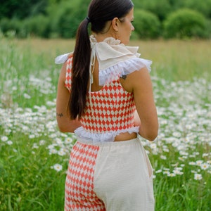 May include: A woman wearing a red and white checkered top with a white lace trim and matching pants. The top is a cropped style and the pants are high-waisted. She is wearing a white lace collar with a bow at the back of her neck. The woman is standing in a field of white daisies.