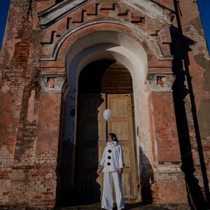 May include: A person in a white outfit with black buttons and a white balloon stands in front of a weathered brick building with a wooden door. The building has a faded pink facade and arched doorway.