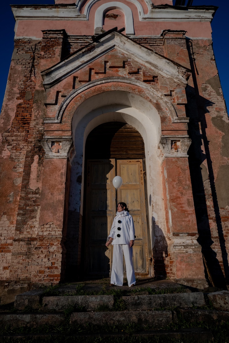 May include: A person in a white outfit with black buttons and a white balloon stands in front of a weathered brick building with a wooden door. The building has a faded pink facade and arched doorway.