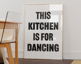 May include: A white framed poster with black text that reads "This Kitchen Is For Dancing". The poster is leaning against a white wall next to a wooden chair.