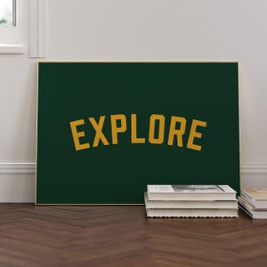 Peut inclure: Une affiche encadrée avec le mot "EXPLORE" en lettres dorées arquées sur un fond vert foncé. Le cadre a une bordure dorée. L'affiche est posée sur un sol en bois, avec une pile de livres devant.