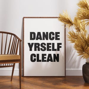 May include: A rectangular wall art print in a dark wood frame featuring the black text DANCE YRSELF CLEAN on a white background. The print is displayed in a room setting with a wooden chair and a dried plant visible to the sides of the frame.