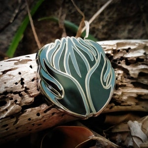 May include: A black and green enamel pin with a stylized design of a hand reaching up with a flame-like shape. The pin has a gold outline.