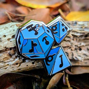 Könnte beinhalten: Ein blau-silberner Emaille-Pin mit drei Würfeln, darunter ein zwanzigseitiger Würfel und zwei kleinere Würfel. Die Würfel haben schwarze Symbole und sind vor einem Hintergrund aus Holz und Blättern platziert. Der Pin hat einen glänzenden silbernen Rand.