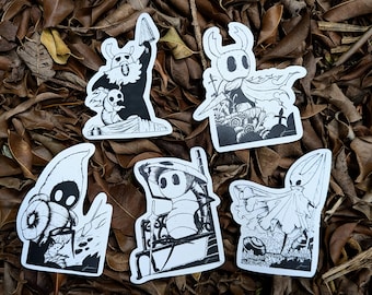 Puede incluir: Cinco pegatinas en blanco y negro con diferentes personajes del videojuego Hollow Knight. Las pegatinas están sobre un fondo de hojas marrones.