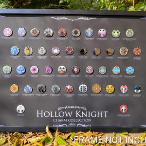 Puede incluir: Una exhibición enmarcada en negro con una colección de pines de encanto Hollow Knight. Los pines circulares tienen varios diseños y colores, dispuestos sobre un fondo oscuro. El texto "Hollow Knight Charm Collection" es visible.