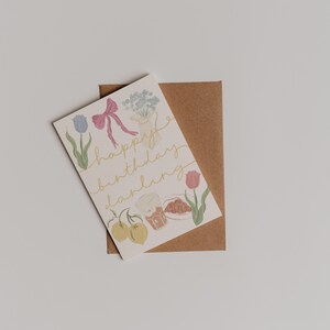 Puede incluir: Una tarjeta blanca con una ilustraci&oacute;n colorida de flores, frutas y un croissant. El texto "happy birthday darling" est&aacute; escrito en letras doradas.