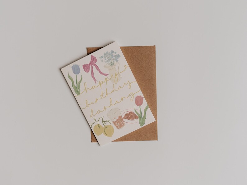 Puede incluir: Una tarjeta blanca con una ilustraci&oacute;n colorida de flores, frutas y un croissant. El texto "happy birthday darling" est&aacute; escrito en letras doradas.