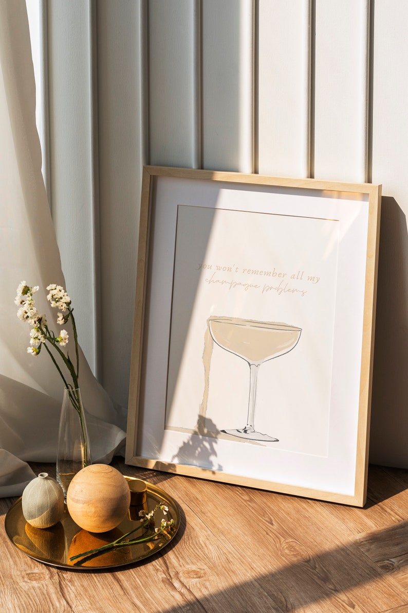 May include: A framed print with a light brown background and a white border. The print features a hand-drawn illustration of a champagne flute with the text "You won't remember all my champagne problems".