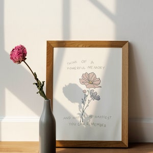 Puede incluir: Una impresión enmarcada con un marco de madera marrón claro y un fondo blanco. La impresión presenta una ilustración dibujada a mano de flores rosas y moradas con el texto "Piensa en un recuerdo poderoso y hazlo el más feliz que puedas recordar."