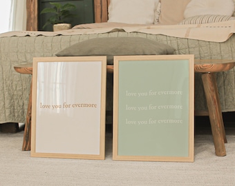 May include: Two framed prints with the text "love you for evermore" in a gold colour. The print on the left has a white background and the print on the right has a light green background. The prints are sitting on a wooden table.