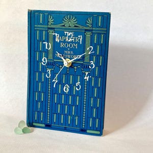 May include: A clock made from a book, titled "The Tapestry Room" by Mrs. Molesworth. The blue cover has a decorative design. The clock face has white numerals and hands, with a gold-coloured centre. The clock is a unique home decor item.