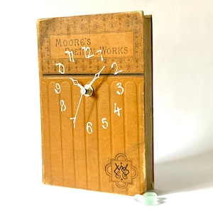 May include: A unique clock made from a vintage book. The clock face is the cover of the book, with the title "Moore's Poetical Works" visible. The clock hands are silver, and the numbers are white. The book is a light brown color.