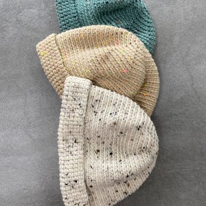 May include: Three knitted beanies in a vertical arrangement. The top beanie is teal, the middle is beige with colourful flecks, and the bottom is off-white with dark flecks. All beanies have a folded brim and a ribbed knit texture.