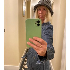 Op de afbeelding: Een vrouw draagt ​​een grijze gebreide bucket hat en een blauw denim jack en maakt een selfie met een groene smartphone.