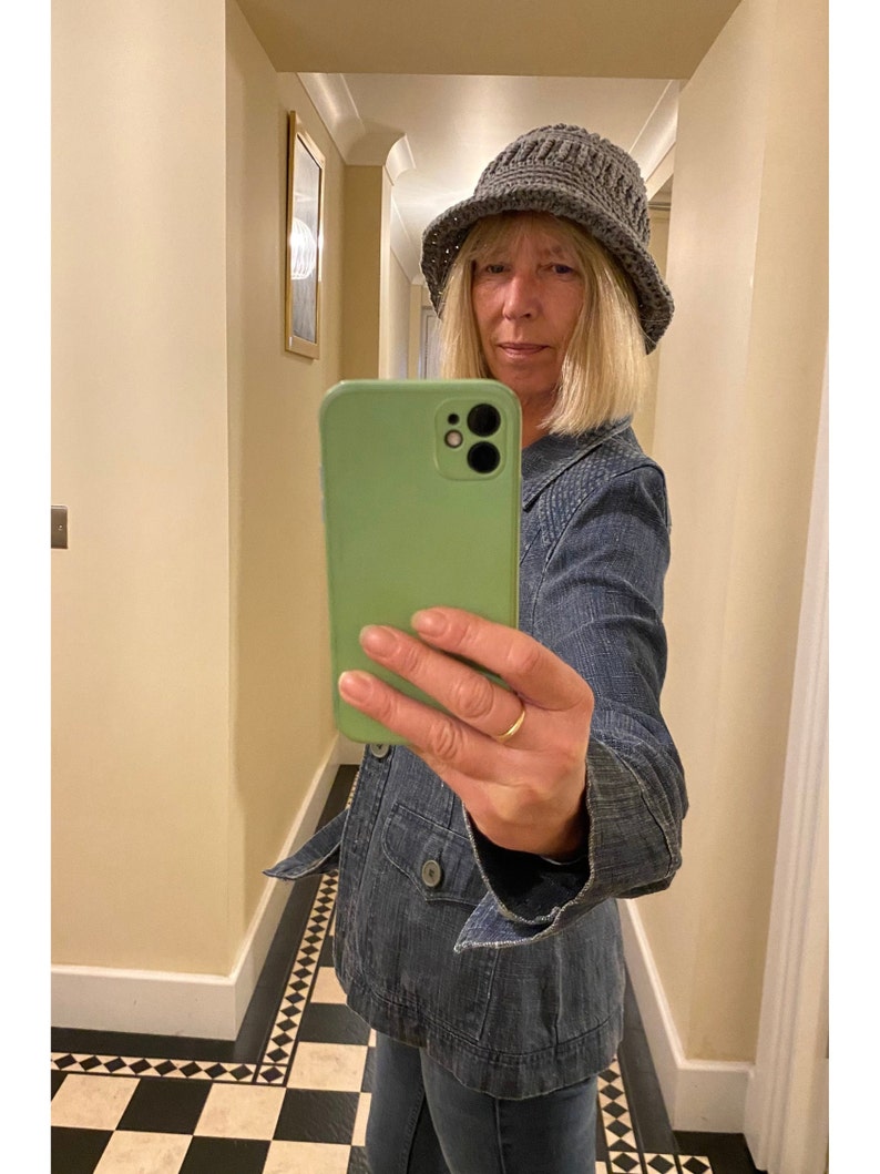 Op de afbeelding: Een vrouw draagt ​​een grijze gebreide bucket hat en een blauw denim jack en maakt een selfie met een groene smartphone.
