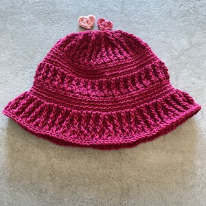 Op de afbeelding: Een roze gebreide bucket hat met twee kleine roze gebreide hartjes erop.