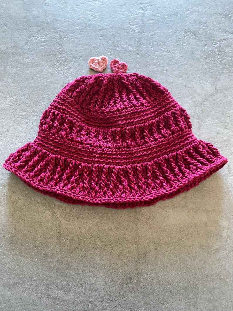 Op de afbeelding: Een roze gebreide bucket hat met twee kleine roze gebreide hartjes erop.