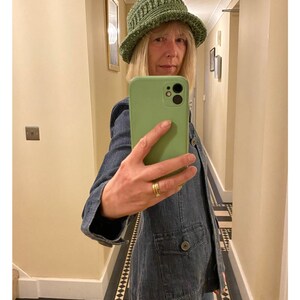 Op de afbeelding: Een vrouw draagt een groene gehaakte bucket hat en een blauw denim jack. Ze maakt een selfie met een groene telefoonhoes.