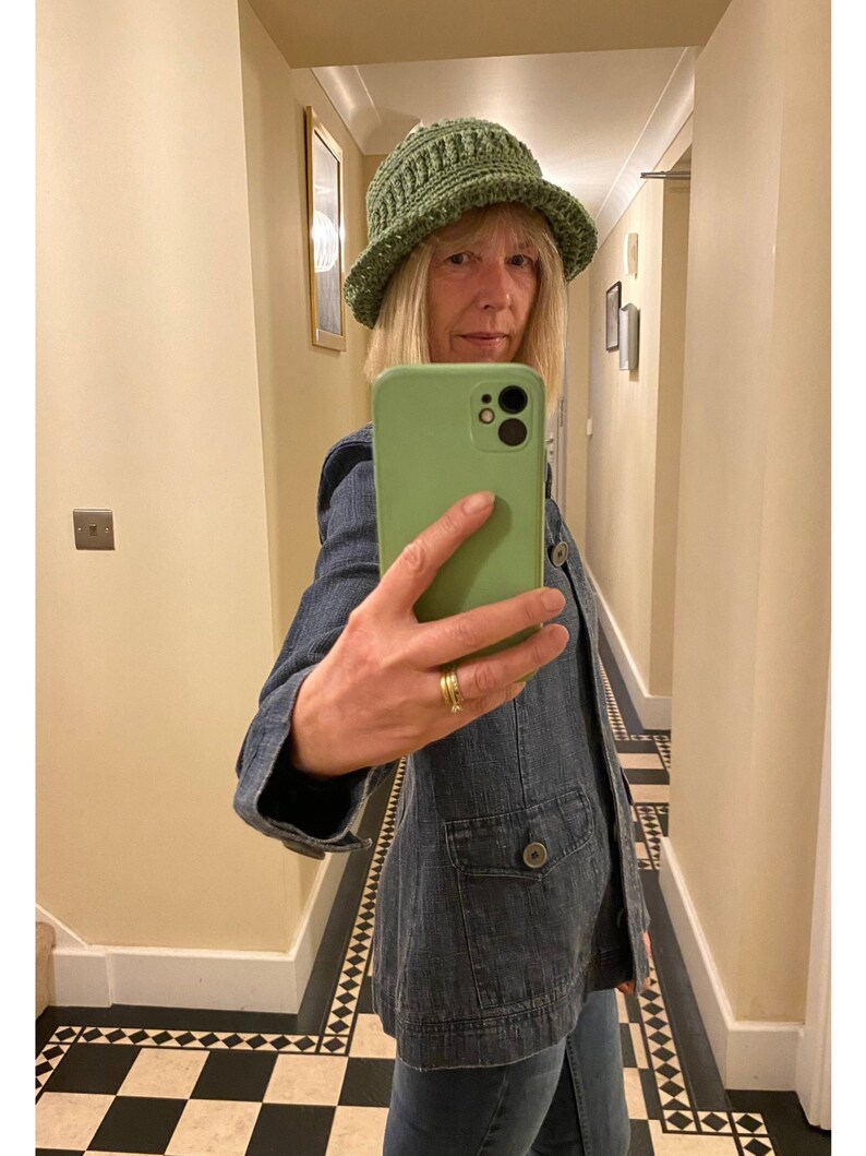 Op de afbeelding: Een vrouw draagt een groene gehaakte bucket hat en een blauw denim jack. Ze maakt een selfie met een groene telefoonhoes.