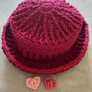 Op de afbeelding: Een bordeauxrode gehaakte hoed met een brede rand en twee kleine roze gehaakte hartjes.