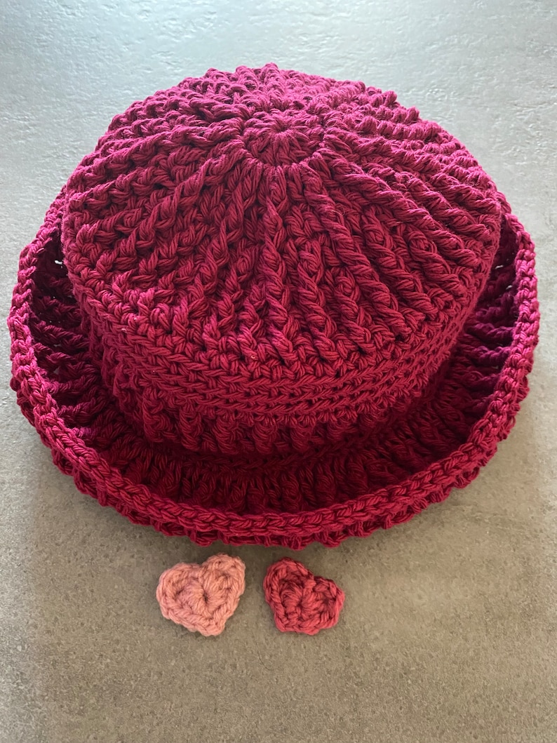 Op de afbeelding: Een bordeauxrode gehaakte hoed met een brede rand en twee kleine roze gehaakte hartjes.