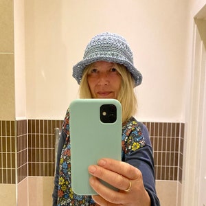 Op de afbeelding: Een vrouw draagt een lichtblauwe gehaakte hoed en een bloemetjesblouse en houdt een lichtblauwe telefoonhoes met een zwarte camera lens vast.