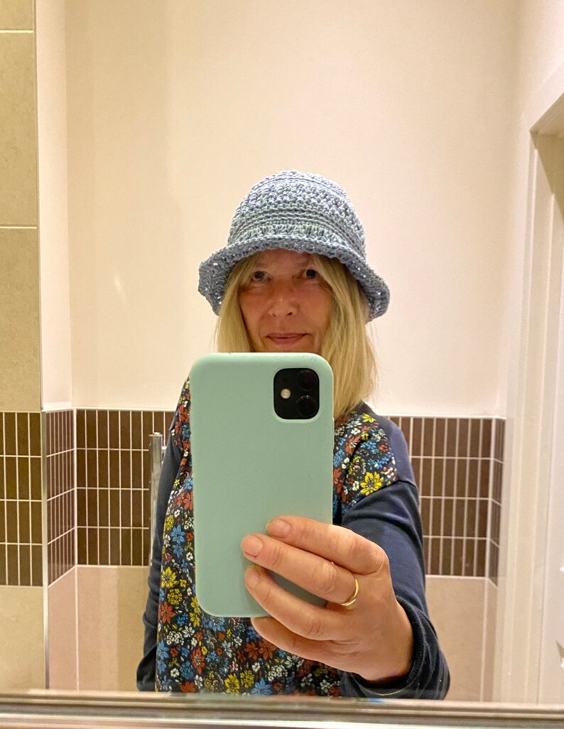 Op de afbeelding: Een vrouw draagt een lichtblauwe gehaakte hoed en een bloemetjesblouse en houdt een lichtblauwe telefoonhoes met een zwarte camera lens vast.