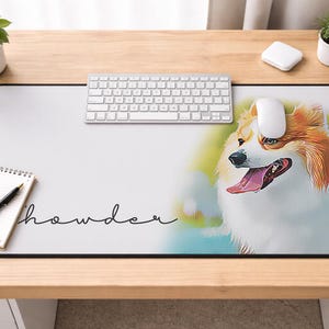May include: A rectangular desk mat with a black border featuring a colorful illustration of a corgi dog with its tongue out. The word "howder" is written in a cursive font. A keyboard, mouse, notepad, and pen are also on the desk.
