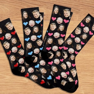 May include: Four black socks with custom faces and heart designs. The socks feature multiple faces and red, pink, and blue hearts. The socks are displayed on a wooden surface.