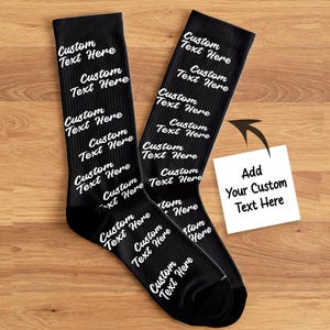 May include: A pair of black socks with the words "Custom Text Here" printed in white. The socks are displayed on a wooden surface. A small white card with the text "Add Your Custom Text Here" is also present.