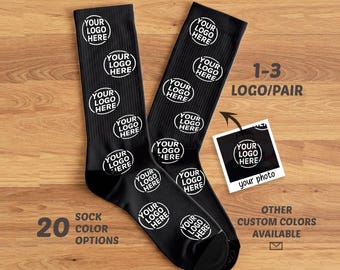 May include: A pair of black crew socks featuring a repeating white circular pattern with the text YOUR LOGO HERE. A small square inset shows a sample logo placement with the text YOUR LOGO HERE and your photo. Text on the image includes 1-3 LOGO/PAIR, 20 SOCK COLOR OPTIONS, and OTHER CUSTOM COLORS AVAILABLE.