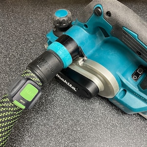 Puede incluir: Una cepilladora eléctrica Makita azul y plateada con una manguera de aspiración de polvo negra. La manguera tiene un botón verde con la etiqueta "MAN".