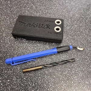 May include: A black foam block with the text "Trackhack" and "32mm 1st Hole" printed on it. The block has two holes drilled into it. A blue and black tool with a curved blade and a gold drill bit are also pictured.