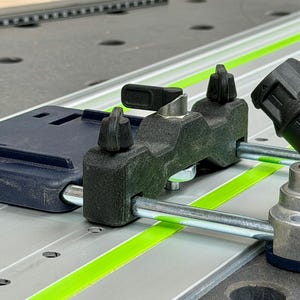 May include: Close-up of a black and silver woodworking tool with a dark blue base. The tool has adjustable knobs and a metal rail with a bright green stripe. The background is a gray work surface.