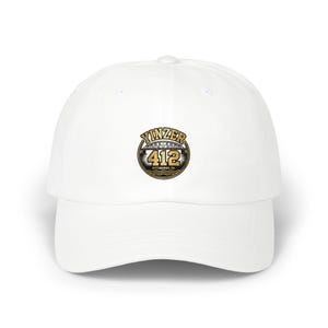 May include: A white baseball cap featuring a circular graphic on the front. The graphic displays the text YINZER in an arched gold font above a bridge illustration, the number 412 in large gold digits, and the text PITTSBURGH, PA below.