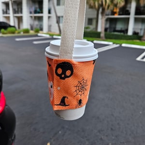 May include: A white to-go coffee cup with a Halloween-themed orange fabric sleeve. The sleeve has a black skull, spider, spiderweb, and witch hat design. The cup is attached to a white fabric strap.