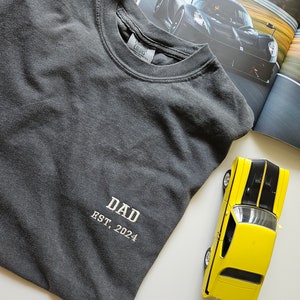 May include: A dark gray t-shirt with the text "DAD EST. 2024" embroidered on the front. The shirt is folded and lying on a white surface.