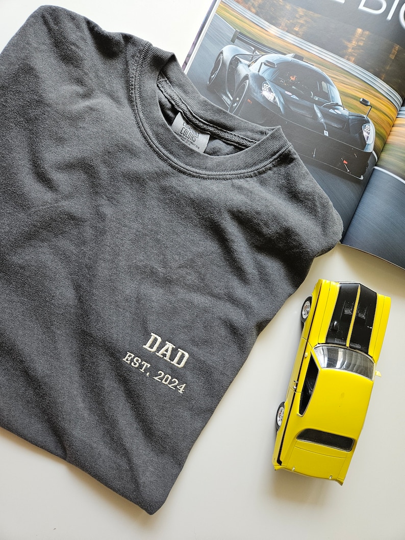 May include: A dark gray t-shirt with the text "DAD EST. 2024" embroidered on the front. The shirt is folded and lying on a white surface.