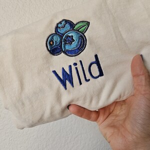 May include: A cream coloured t-shirt with an embroidered design of three blue blueberries and a green leaf. The word "Wild" is embroidered in blue below the blueberries.