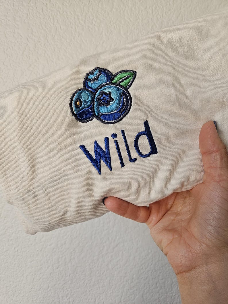 May include: A cream coloured t-shirt with an embroidered design of three blue blueberries and a green leaf. The word "Wild" is embroidered in blue below the blueberries.