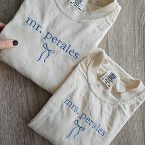 May include: Two cream-colored t-shirts with blue embroidered text and bow details. One shirt reads "mr. perales" and the other reads "mrs. perales". Both shirts have a Comfort Colors tag. The shirts are laid on a wooden surface.