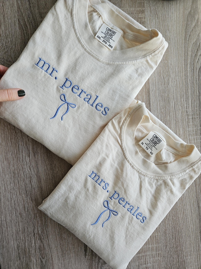 May include: Two cream-colored t-shirts with blue embroidered text and bow details. One shirt reads "mr. perales" and the other reads "mrs. perales". Both shirts have a Comfort Colors tag. The shirts are laid on a wooden surface.