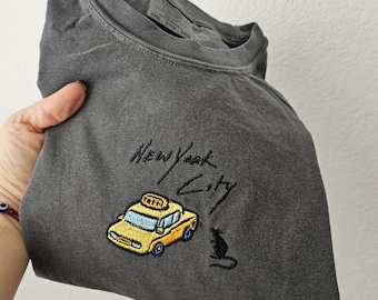 May include: Dark gray t-shirt with embroidered design. Features a yellow taxi cab with blue accents and the words "New York City" in script. A small black cat is also embroidered on the shirt.