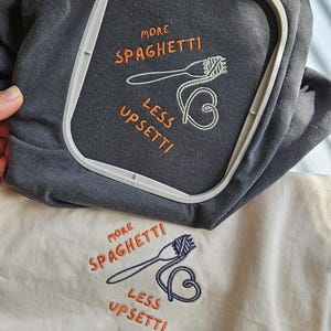 May include: Two embroidered t-shirts with the text "More Spaghetti Less Upsetti" in orange and a white embroidered image of a fork and spaghetti. The t-shirts are one gray and one cream.