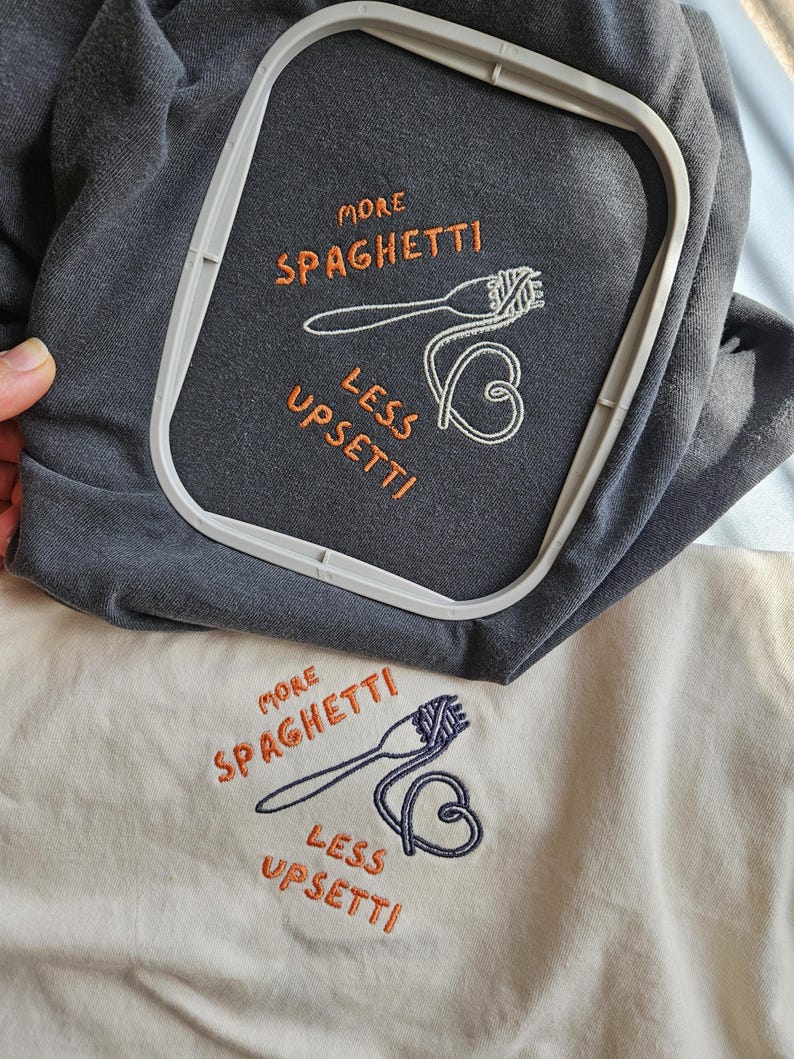 May include: Two embroidered t-shirts with the text "More Spaghetti Less Upsetti" in orange and a white embroidered image of a fork and spaghetti. The t-shirts are one gray and one cream.